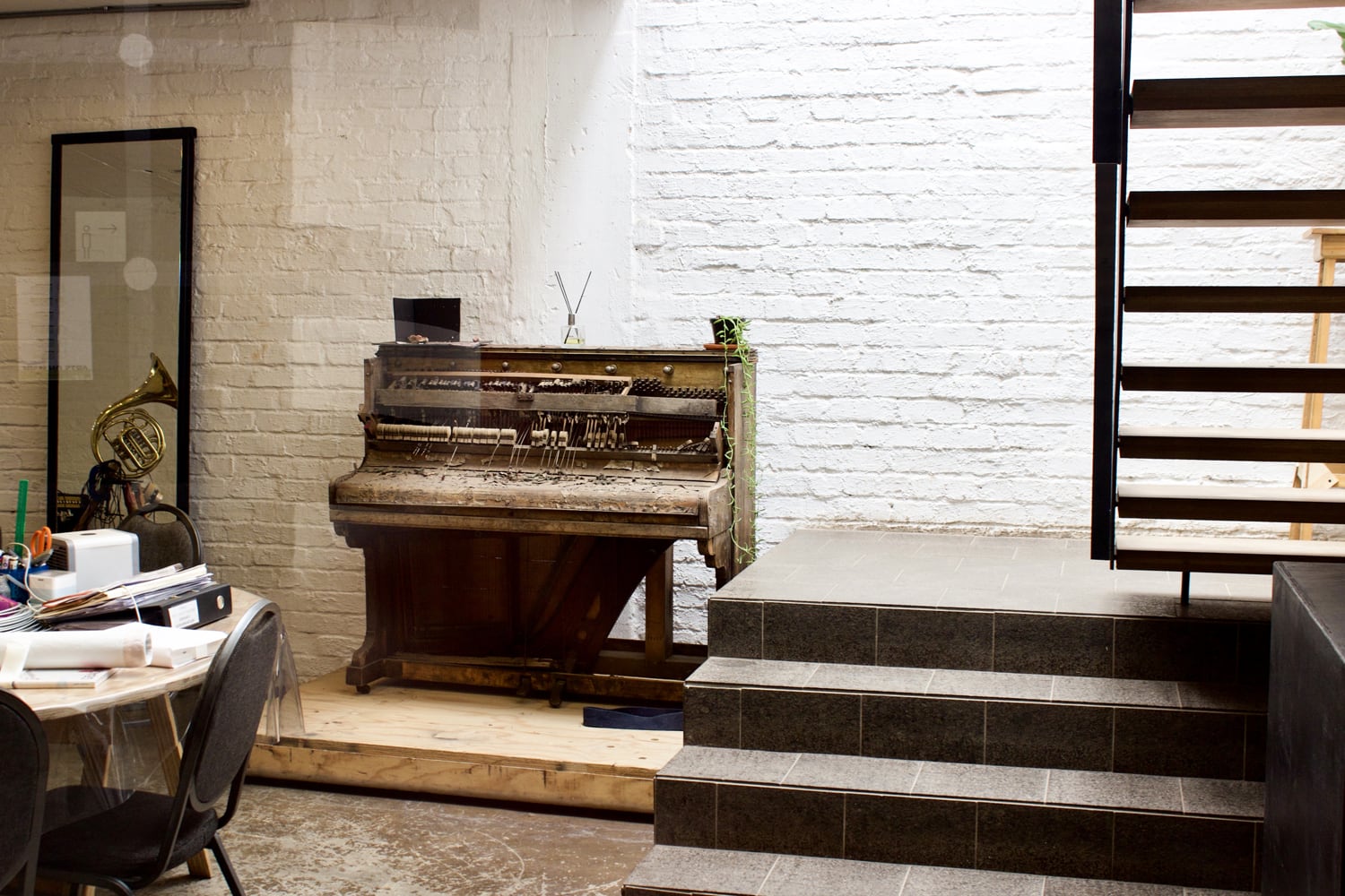 An upright piano with exposed interior stands on a wooden platform against a white brick wall. To the left, a French horn rests behind a chair and cluttered table. Modern stairs with grey tiles rise on the right side of the image.