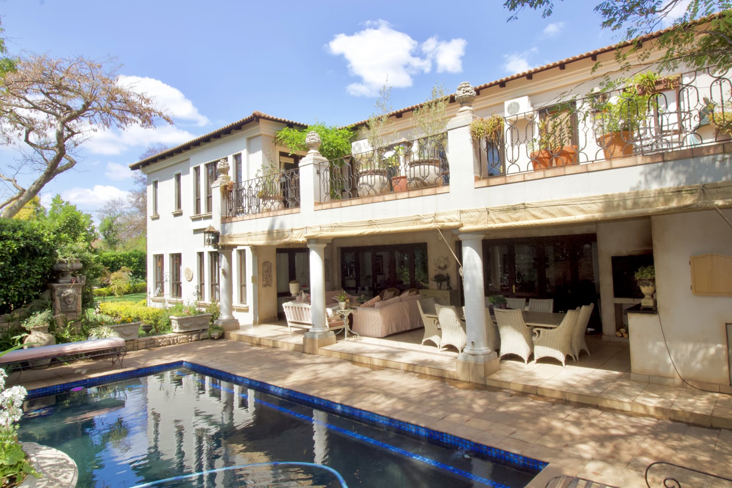 Home 3 A two-storey Mediterranean-style house with a balcony and a covered patio furnished with wicker chairs and sofas. The outdoor area features a rectangular swimming pool bordered by stone tiles, surrounded by lush greenery and trees under a bright blue sky with clouds.