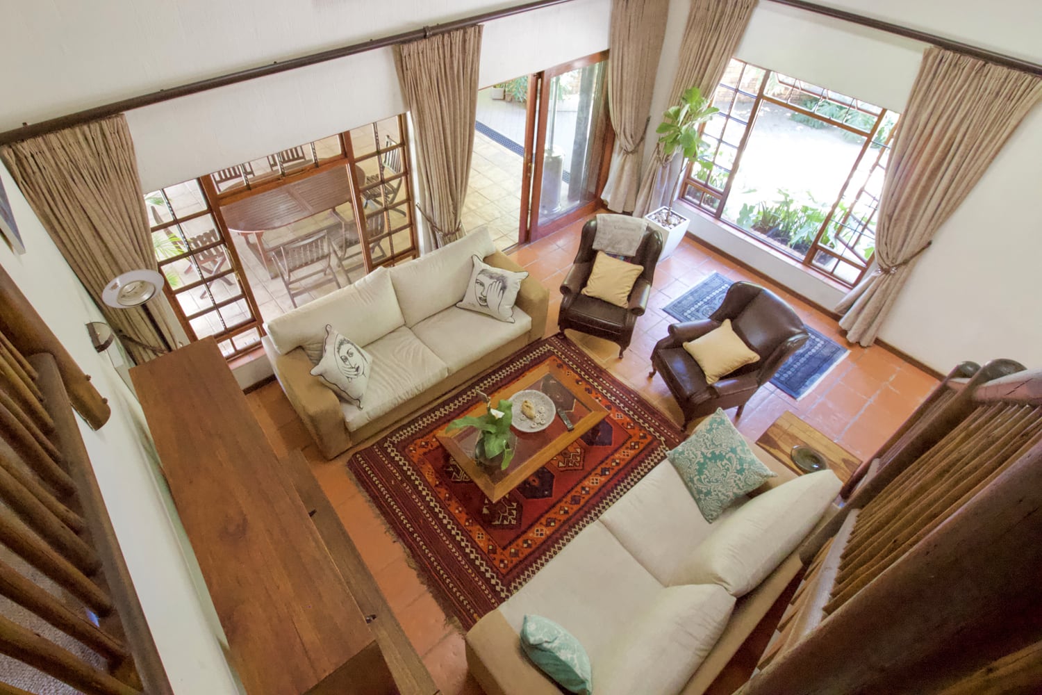 Home 1 View of a cosy living room from above, featuring two beige sofas and two armchairs around a wooden coffee table on a patterned red rug. Large windows with beige curtains let in natural light. There are plants, cushions, and a wooden cabinet in the room.