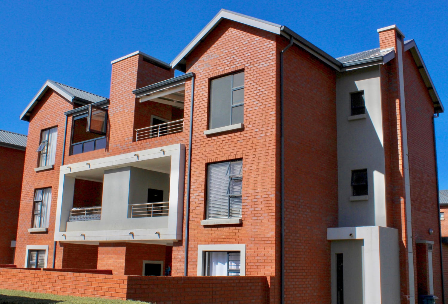 A modern, three-storey block of flats with red brick walls and grey accents. It features two covered balconies, multiple large windows, and a flat roof. The sky is clear and blue, and a low brick wall borders the front of the building.