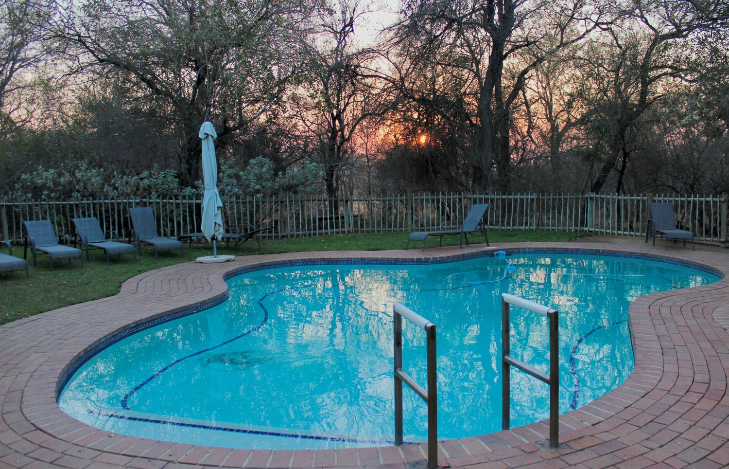 A clear blue swimming pool with metal handrails is surrounded by a brick terrace, loungers, and a parasol. Trees and a fence enclose the area, and the sun sets in the background, casting a warm glow through the branches.