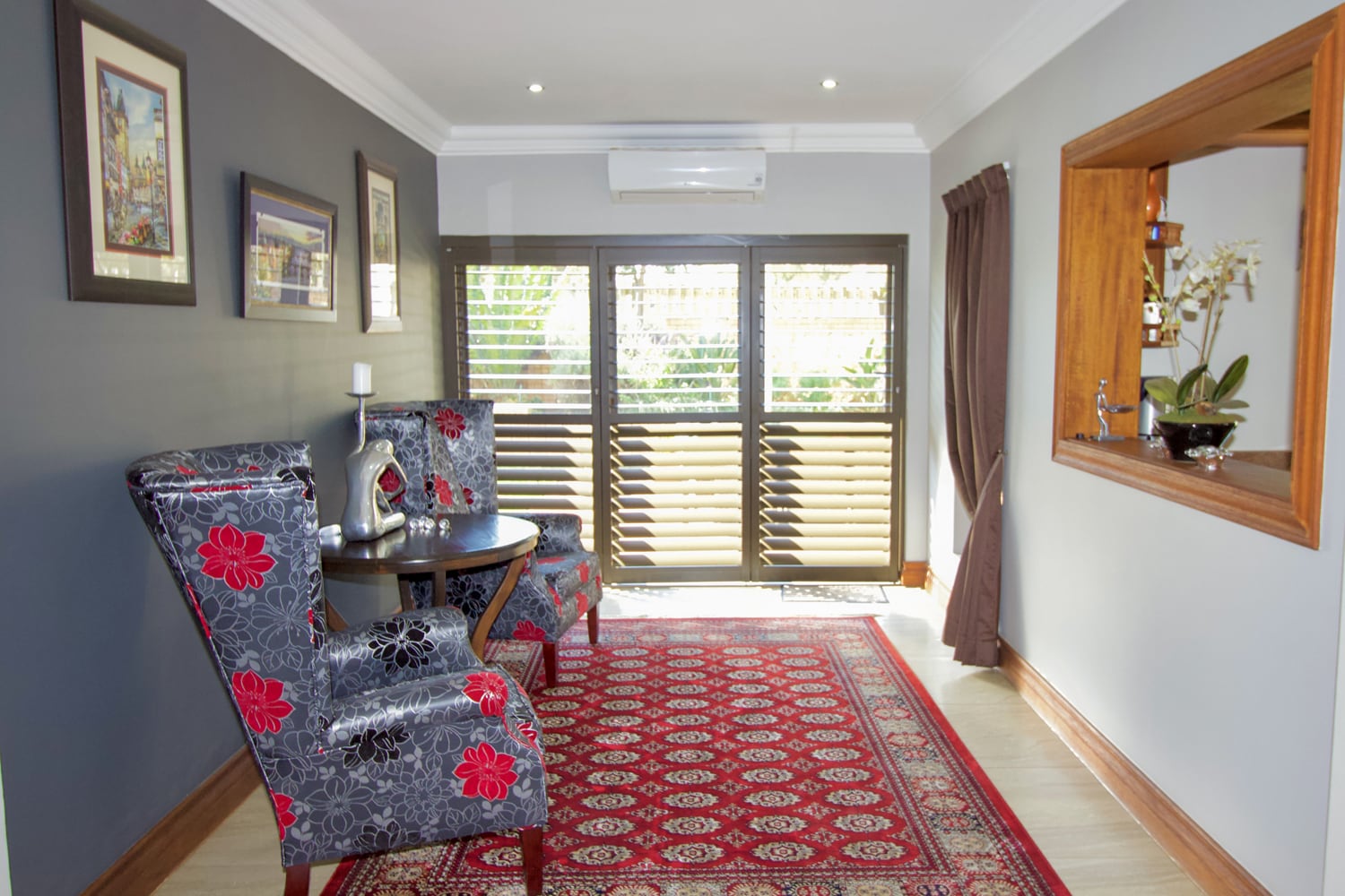 Small sitting area with two floral-upholstered chairs, a round table, and a red patterned rug. Grey feature wall with framed art, large windows with wooden shutters, indoor plant on a shelf, and an air conditioner above the windows.