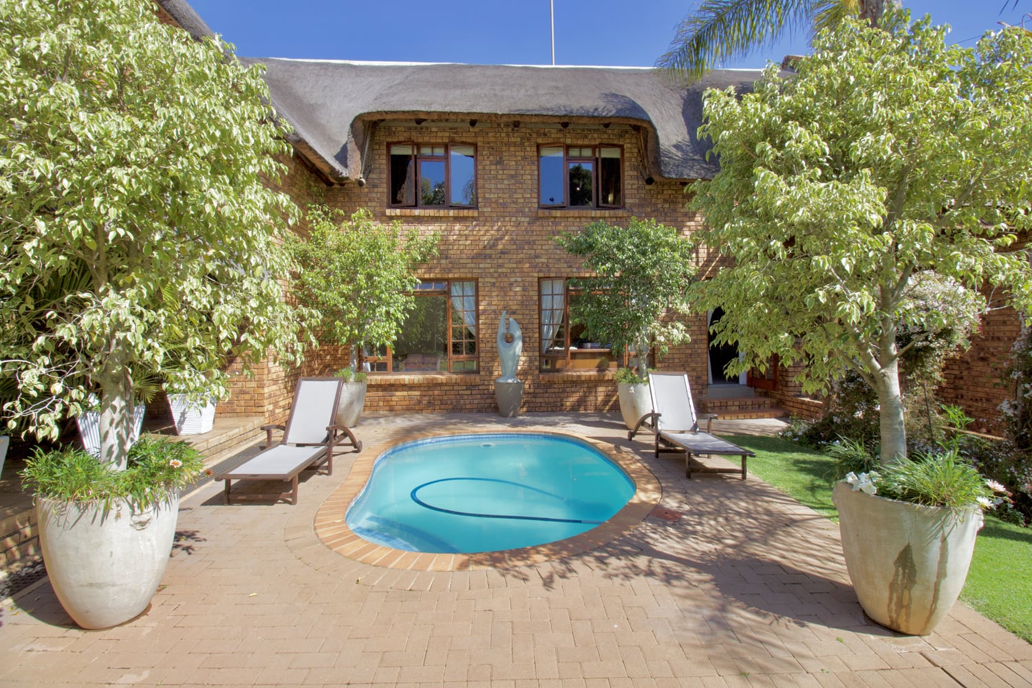 A small, oval swimming pool sits in a brick patio surrounded by potted plants and trees. Four sun loungers line the poolside. In the background is a two-storey brick house with a thatched roof, large windows, and a central garden statue.