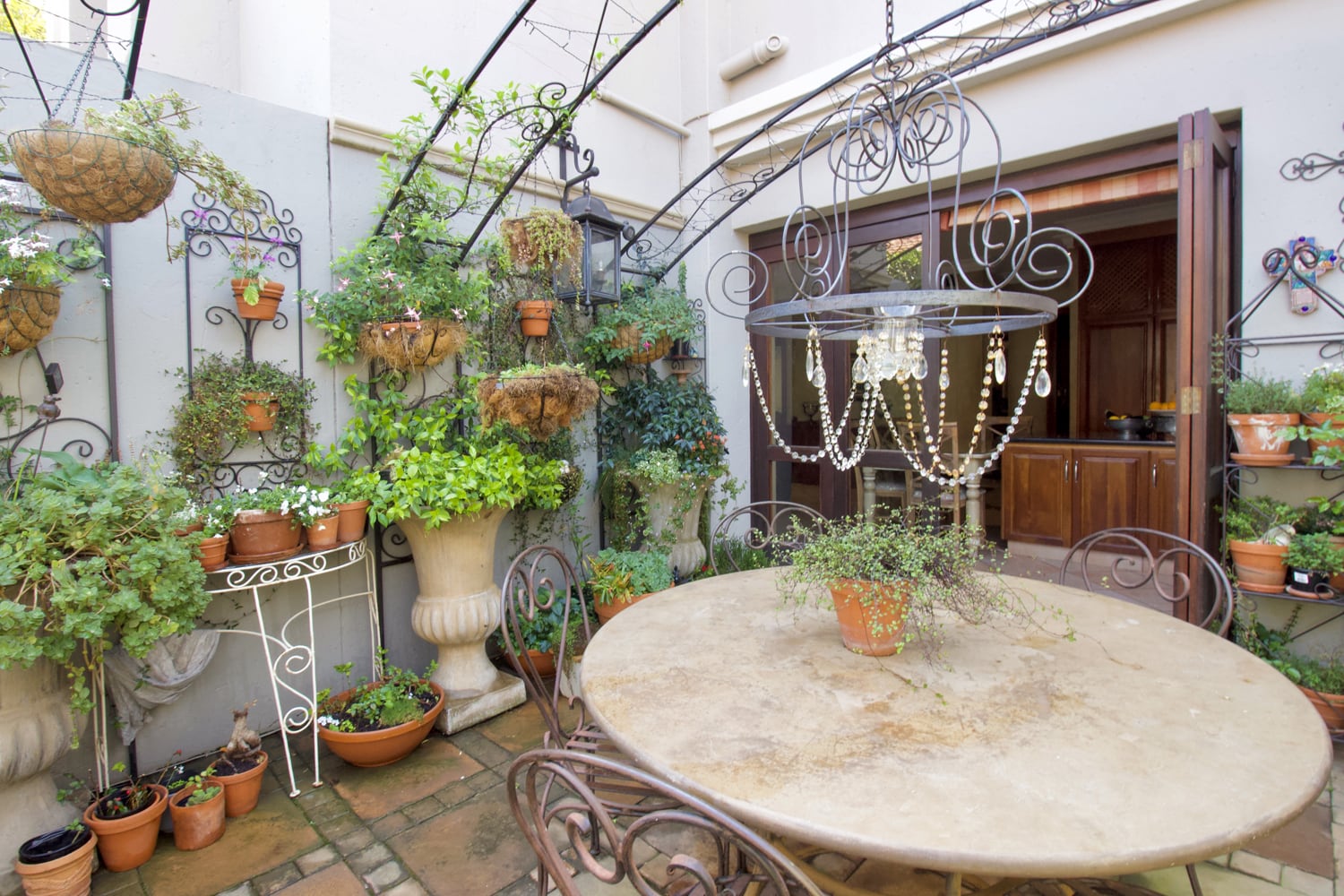 An outdoor patio with a round stone table and metal chairs, a beaded chandelier, many potted plants on shelves and hanging baskets, and a view into a kitchen through an open door. The area is bright and decorated with greenery.