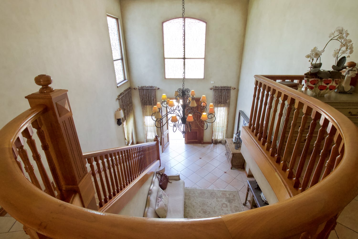 View from the top of a curved wooden staircase overlooking a bright foyer with tiled floor, large arched window, chandelier, beige walls, and a sitting area with a sofa, rug, and decorative side tables. Sunlight streams in through the windows.