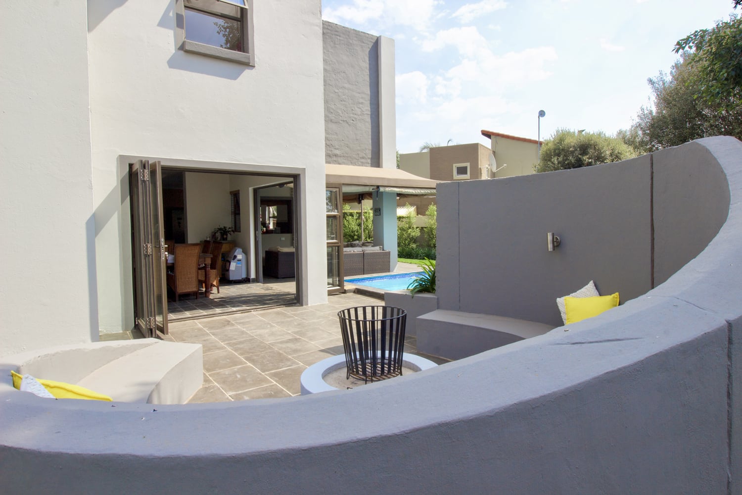 Modern outdoor patio with curved grey seating around a circular fire pit, accented by yellow cushions. Sliding glass doors lead to a contemporary house interior. A small pool and greenery are visible in the background under a partly cloudy sky.