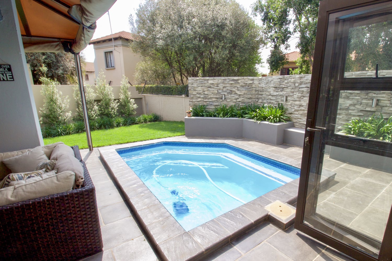 A small, modern back garden pool with blue water is surrounded by grey stone tiles. A cushioned patio sofa sits on the left. Green grass, shrubs, and a stone wall with planters are in the background. A glass door is open at the pool’s edge.