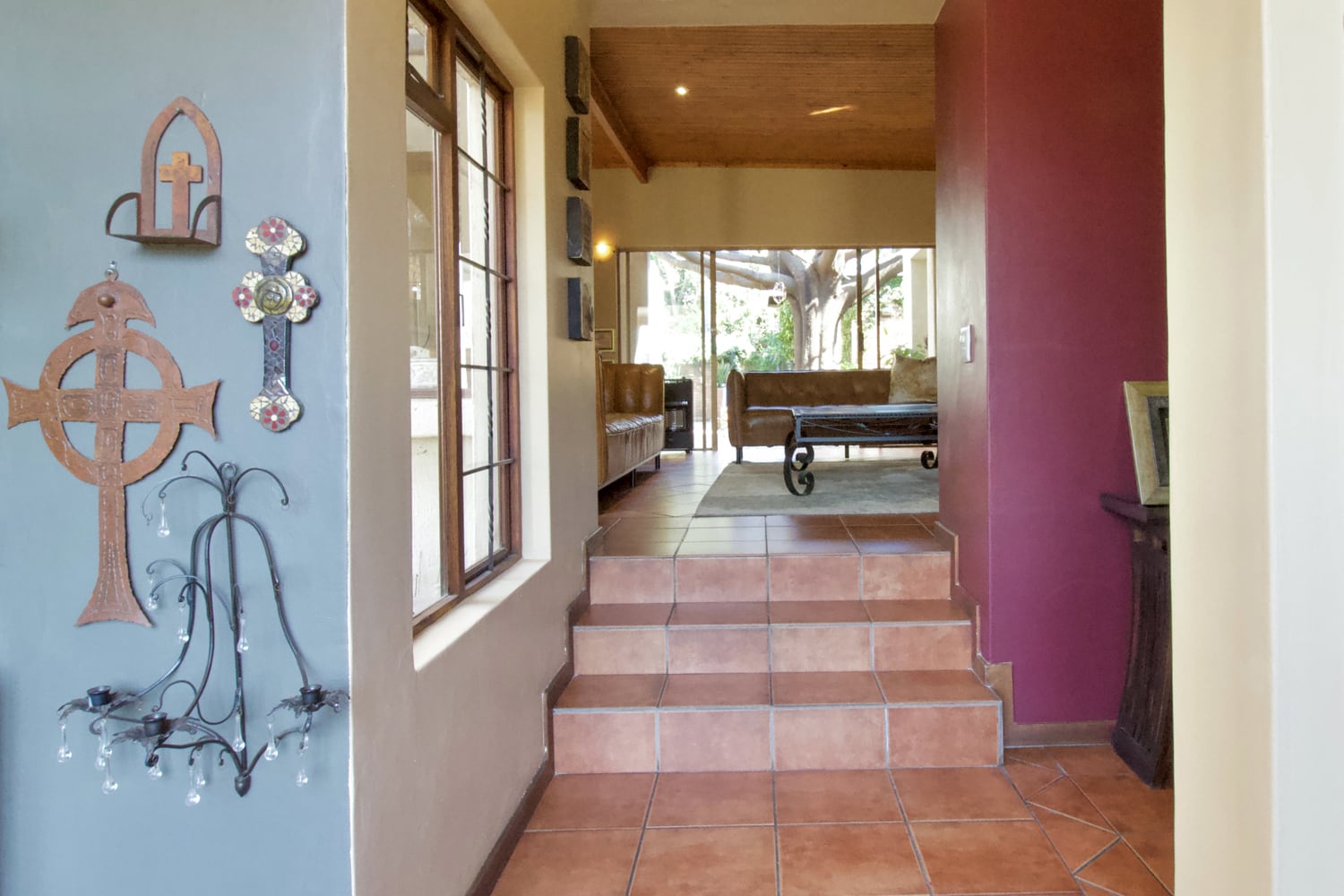 A home interior with terracotta tile floors and a few steps leading to a living area. The left wall has decorative crosses, while large windows let in natural light. The living space has sofas and a view of a tree outside. Walls are painted beige, blue, and maroon.